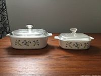 Two white ceramic Corning Ware casserole dishes with floral patterns on the side, each with a glass lid, displayed side by side on a wooden surface.