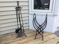 Matt black heavy iron fireplace tool set with stand holding poker, brush, shovel, and tongs. Curved heavy iron firewood log holder beside it.