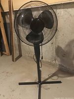 Photo shows a black 42-inch floor fan with three blades and a metal grille on an adjustable height stand with a T-shaped base placed on a concrete floor.
