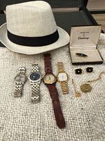 Photo of four men's wristwatches, a beige fedora hat, vintage cuff links, tie clips, and gold-tone jewelry on a textured surface.