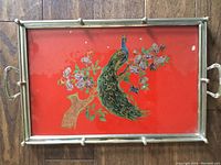 Front view of the peacock tray showing the red background with peacock and floral design and silver metal framed edges with handles on both sides.