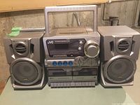 Front view of JVC portable stereo system showing main unit with CD player, radio controls, tape deck, and two detachable speakers.