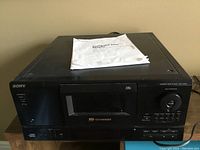 Front view of Sony 100 CD changer player showing control buttons, disc chamber and power switch. Original operating manual placed on top.