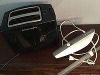 KitchenAid black two-slot toaster placed next to Hamilton Beach electric cutting utensil with visible power cord and protective blade sheath on wooden surface.