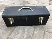 Closed black metal tool box with white handle and silver latches on a paved surface