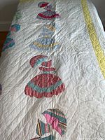 Full view of antique handmade quilt laid on a bed showing colorful women figures with umbrellas appliquéd onto a white base fabric, with yellow patterned border.
