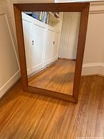 Mirror standing on hardwood floor against white cabinet background showing the full wood frame and reflective glass surface.