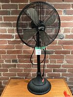 Full view of black metal standing fan with adjustable height and round base against brick wall.