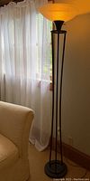 Full view of the antique style torchiere floor lamp next to a window with sheer curtain and beige couch, showing bronze metal frame and lit frosted glass shade.