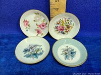Photo of four Royal Worcester small butter pat dishes with various floral decorations and gold rims on blue background with ruler for size reference.