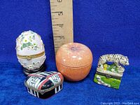 Lot of four assorted decorative trinket boxes: white floral egg-shaped box on stand, peach-shaped box, tiger figurine box, piano-themed box with musical notes and colors