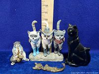 Group of six cat-themed figurines including a ceramic saxophone-playing cat, three cowboy-dressed cats, a black reclining cat, a bronze cat lying down, and the black cat plant pot.