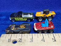 Four diecast toy vehicles displayed on blue background with a ruler measuring roughly 2-3 inches in length each.
