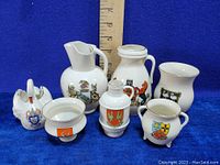 Full view of all 6 miniature Goss Ware porcelain pieces with various English crests and coats of arms on a blue background with a measuring stick for scale