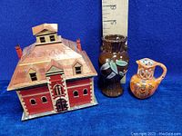 Photo of the house-shaped ceramic box in reddish-brown with brick and roof details, alongside brown vase with green leaf decoration and small orange jug