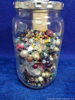 Clear glass jar filled to brim with multi-colored assorted jewelry beads, viewed from side.