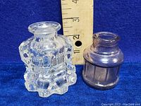 Photo showing clear crystal cut glass ink bottle alongside an amethyst-colored glass ink bottle with a ruler for size reference, both approximately 2 and 1.5 inches tall respectively.