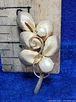 Close-up view of vintage flower brooch with two faux pearls and textured silver leaves on a wooden ruler background for size reference.
