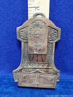 Front view of the vintage coppered tin plate match box holder and striker showing embossed decorative elements and hook for wall hanging.