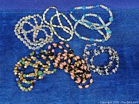 Lot of glass bead necklaces arranged on a blue velvet surface, showing variety of colors and styles including clear, pink, black with gold, and blue beads.