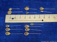 View of all ten gold tone filigree hat and scarf pins arranged on a blue background with ruler for scale demonstrating lengths from about 2 to 3.5 inches.