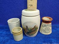 Photo showing three miniature crocks with a measuring stick for size reference against a blue background. Includes the James Nicoll Scotland ginger jar with bird design, the unsigned small white crock, and the Lancashire Dairies cream crock.