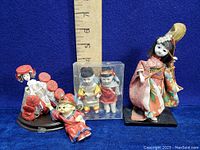 Photo showing multiple Asian porcelain dolls and figurines with a ruler for size reference, featuring a Japanese kimono doll, small children figures, and other colorful figurines.