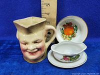 Photo of a vintage happy face mug with two small dishes, one with fruit design and one with floral design, showing size with ruler.