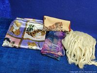 Four scarves and wraps displayed on blue background. One scarf with various cat prints and lavender frame, one leopard print scarf, one purple-blue batik-style scarf, and one cream mohair fringed shawl wrap.