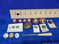 Multiple buttons arranged with ruler for size; features red floral glass buttons, colorful glass jewel button, mother of pearl buttons, metal globe button, and mother of pearl button hook.