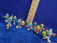 Full set of 12 vintage mini Smurf vinyl figurines arranged on blue background with ruler for size reference showing measurements about 1.5-2 inches tall each.