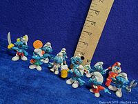 Nine vintage mini Smurf vinyl figures posed in a line on blue background with ruler to show size around 2-3 inches tall.
