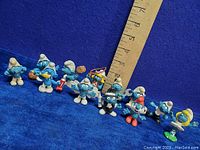 Photo showing 11 vintage Smurf vinyl figures posed with a ruler measuring approximately 5 inches tall, displaying various characters in different poses and holding accessories.