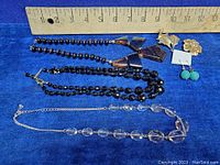 Photo showing three vintage necklaces laid out against a blue background with a ruler for size reference. Necklaces include black glass beads, clear glass beads, and dark beads with metal cone and geometric accents.