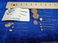 Photo showing full lot of 14 vintage hat and scarf pins arranged on blue fabric with ruler for scale. Includes pins with gold filigree and faux pearl tops in various shapes and sizes.