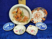 Full lot showing two large decorative plates, two small floral salt cellars, and two blue and white small oval dishes on blue fabric background with ruler for scale.