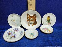 Six decorative plates arranged on blue cloth backdrop with size scale showing approx 4 to 7 inches diameter.