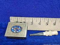 Photo showing the metal matchbox with detailed floral engraving and blue enamel floral oval insert, alongside the carved ivory holder next to a ruler for scale.