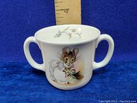 Front view of white porcelain child's mug with two handles, decorated with Hunca Munca mouse illustration holding a broom and dustpan. Small floral design inside rim. Blue background and wooden ruler for scale.