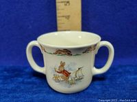 Front side of the white ceramic double handled child's mug featuring a bunny in a red sweater sailing a small boat with a duckling, with a border of running rabbits above.