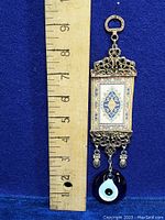 Full-length view of the wall hanging next to a ruler showing it is nearly 7.5 inches long, featuring the evil eye glass at the bottom.