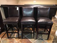 Three bar stools with dark brown wood legs and dark brown vinyl cushioned seats and backs arranged side by side on tiled floor.