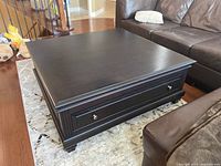 Full view of black laminated coffee table with dovetailed front drawer, sitting on patterned rug next to leather sofas
