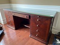 Front view of the desk showing wood laminate finish, glass top, multiple drawers, and cabinet doors.
