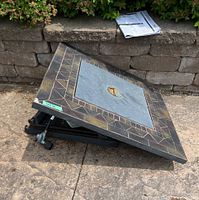 Top view of the square mosaic fire table top with decorative tile edge and central metal cover with valve knob, rest on black metal frame parts outdoors. Instruction sheet partially shown on stone wall.