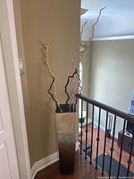 Full view of tall square floor vase with twisted decorative branches inside, placed next to railing indoors.