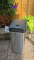 Photo of the iTouchless outdoor garbage pail positioned in outdoor garden setting showing stainless steel body and black plastic top lid with touchless controls.