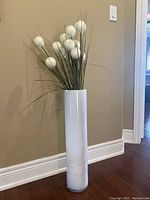 Tall white cylindrical glass vase with faux flower arrangement inside, located on hardwood floor against beige wall.