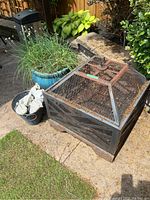 Outdoor photo showing the rusted metal framed fire pit with geometric cutouts on the sides and wire mesh cover on top. Next to it is a metal bucket containing wrapped fire logs.