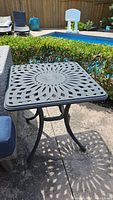 Outdoor cast metal side table square top showing sunburst lattice design, placed patio next to blue and gray wicker sofa.